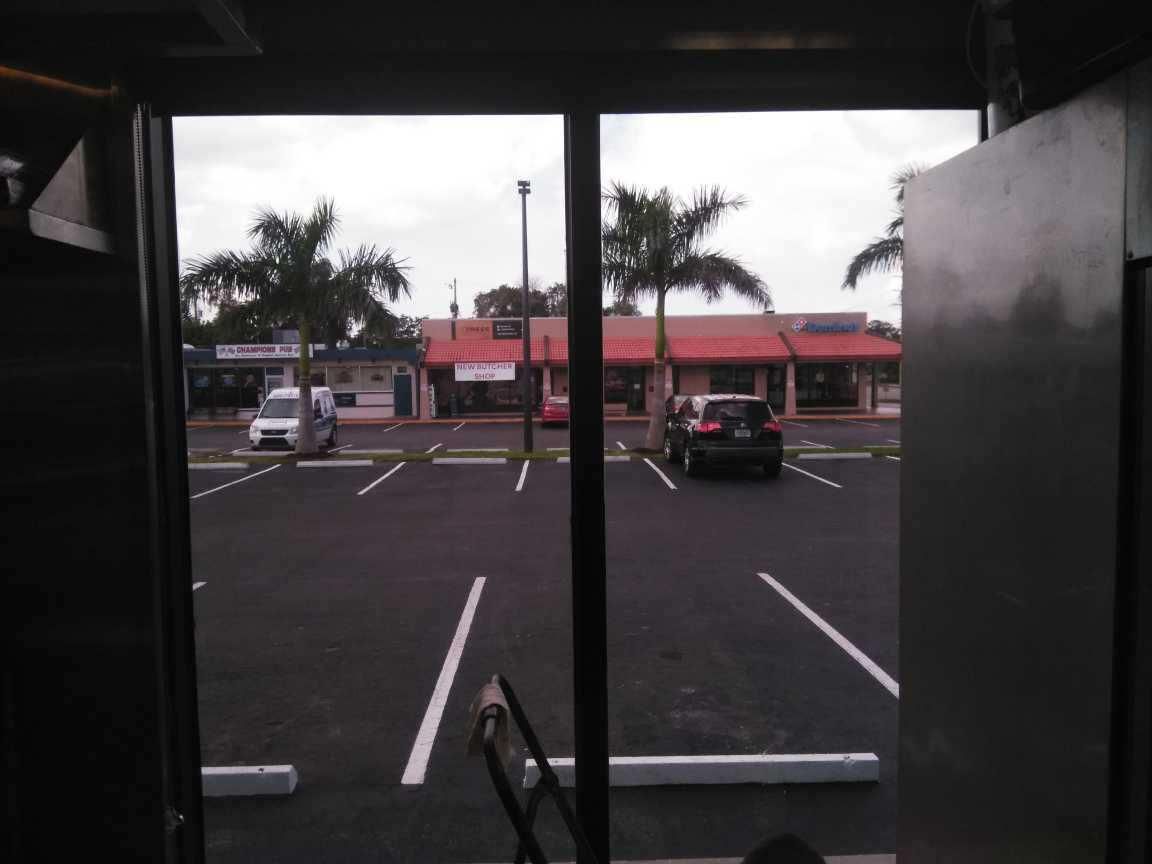 View from a covered parking lot toward a red-roofed storefront and palm trees on a cloudy day