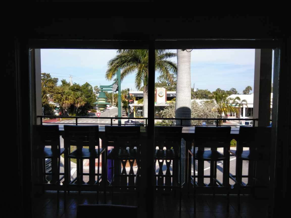 View through a dark balcony railing toward a sunny street with palm trees and buildings.