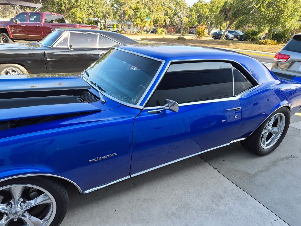 Blue classic muscle car parked in a driveway, side view with black roof and chrome wheels.
