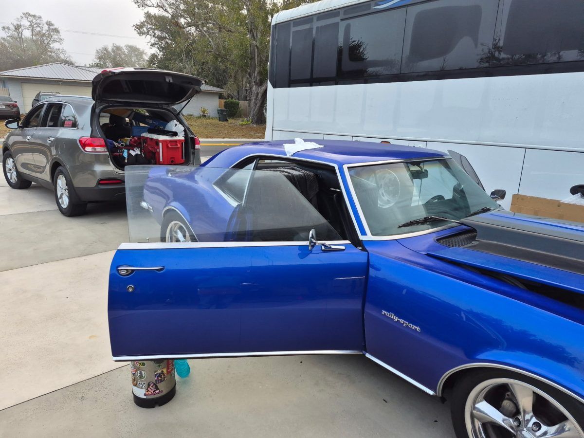 Blue classic car with open door and hood parked beside a white bus in a driveway