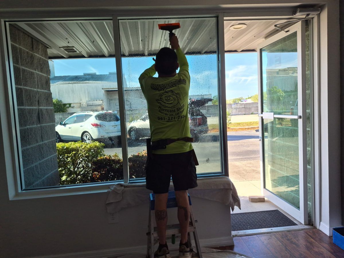 Person on a ladder painting the top of a large window inside a room.