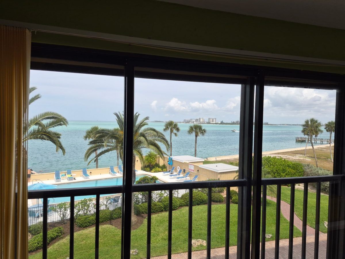 Ocean view from a balcony with palm trees, pool, beach umbrellas, and grassy lawn.