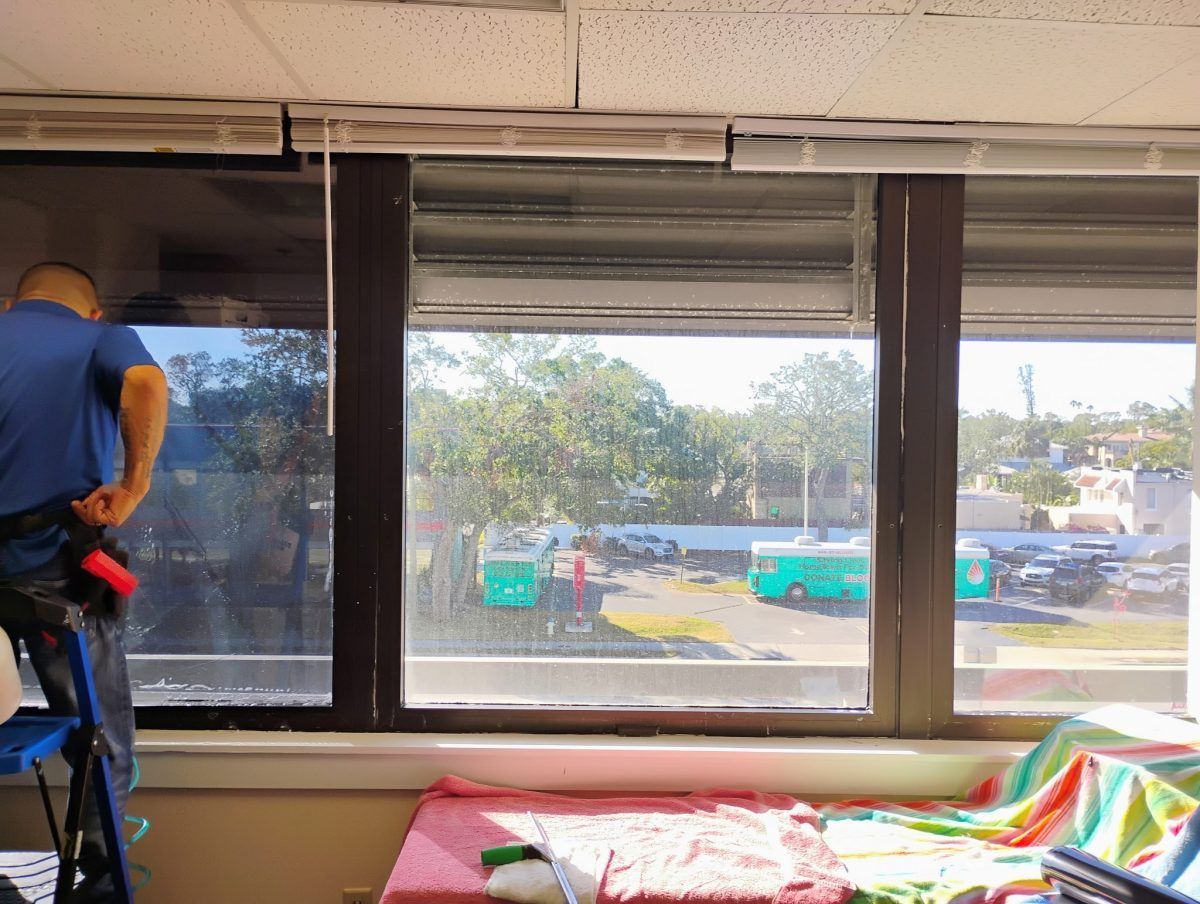Worker cleaning windows beside a bed in a room with large windows and a colorful blanket
