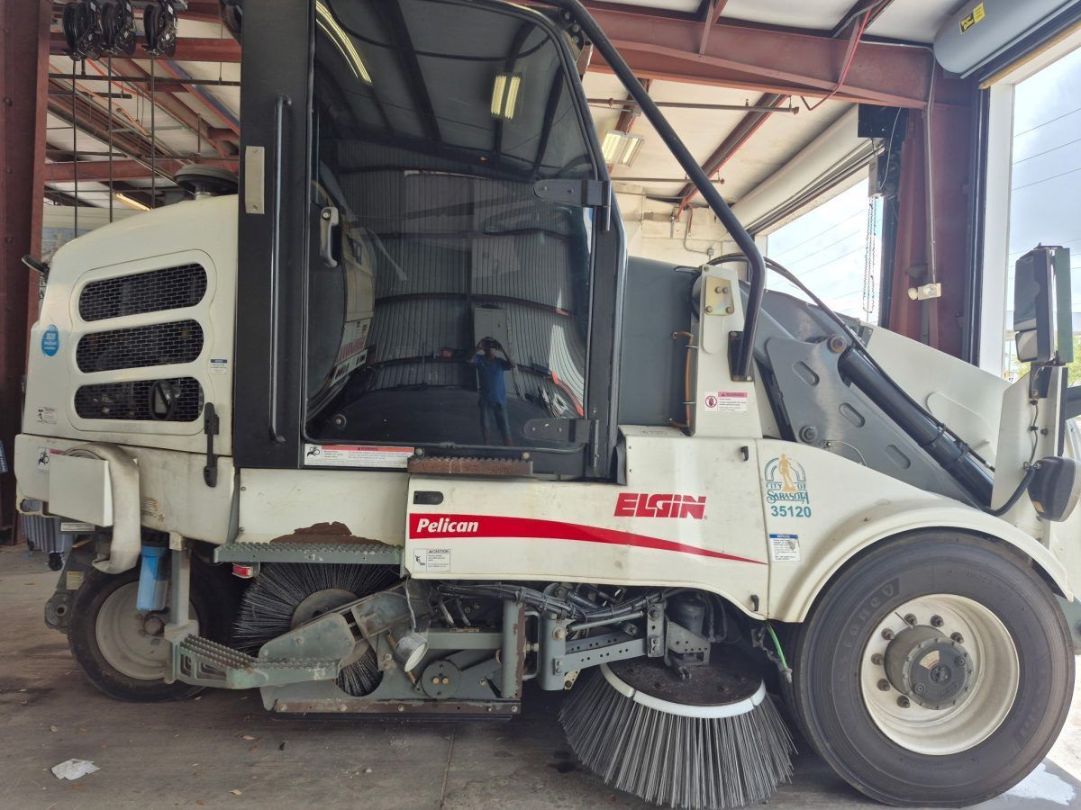 White street sweeper parked in a warehouse, showing front brush and side vents