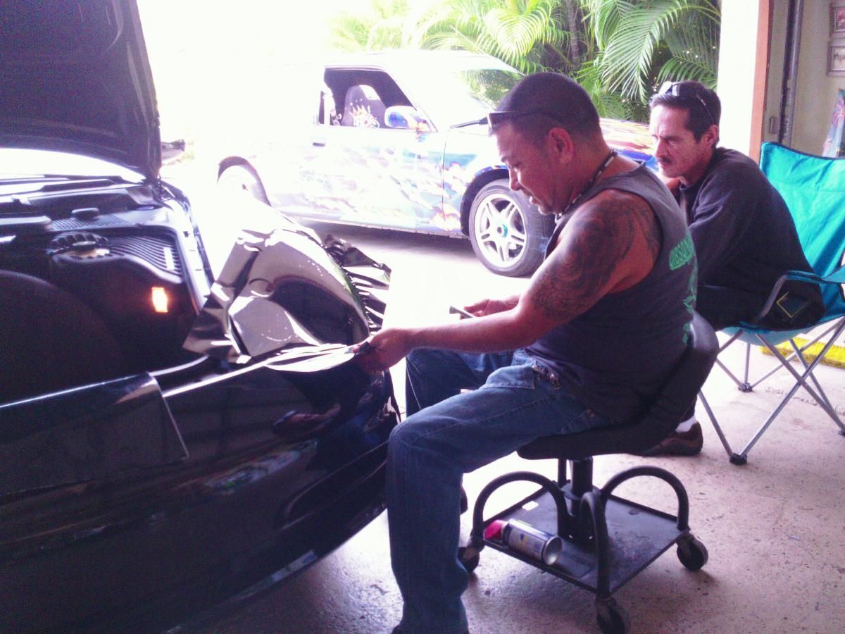 Mechanic inspecting a car engine with hood open in a garage, seated on a rolling stool.