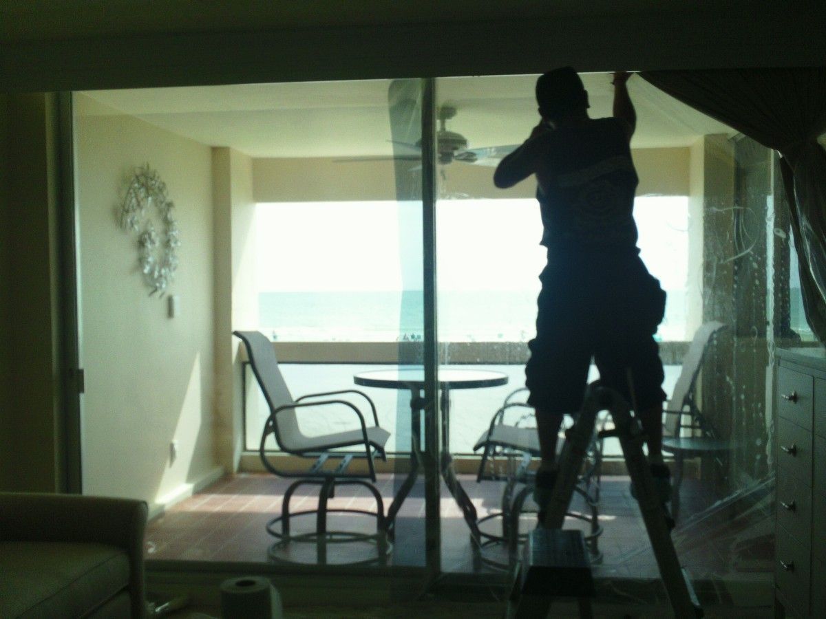 Person standing on a step ladder cleaning a glass door in a bright room with patio furniture.