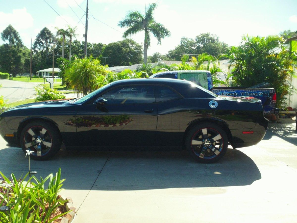Black sports coupe parked in a sunny driveway beside palm trees and a blue pickup truck