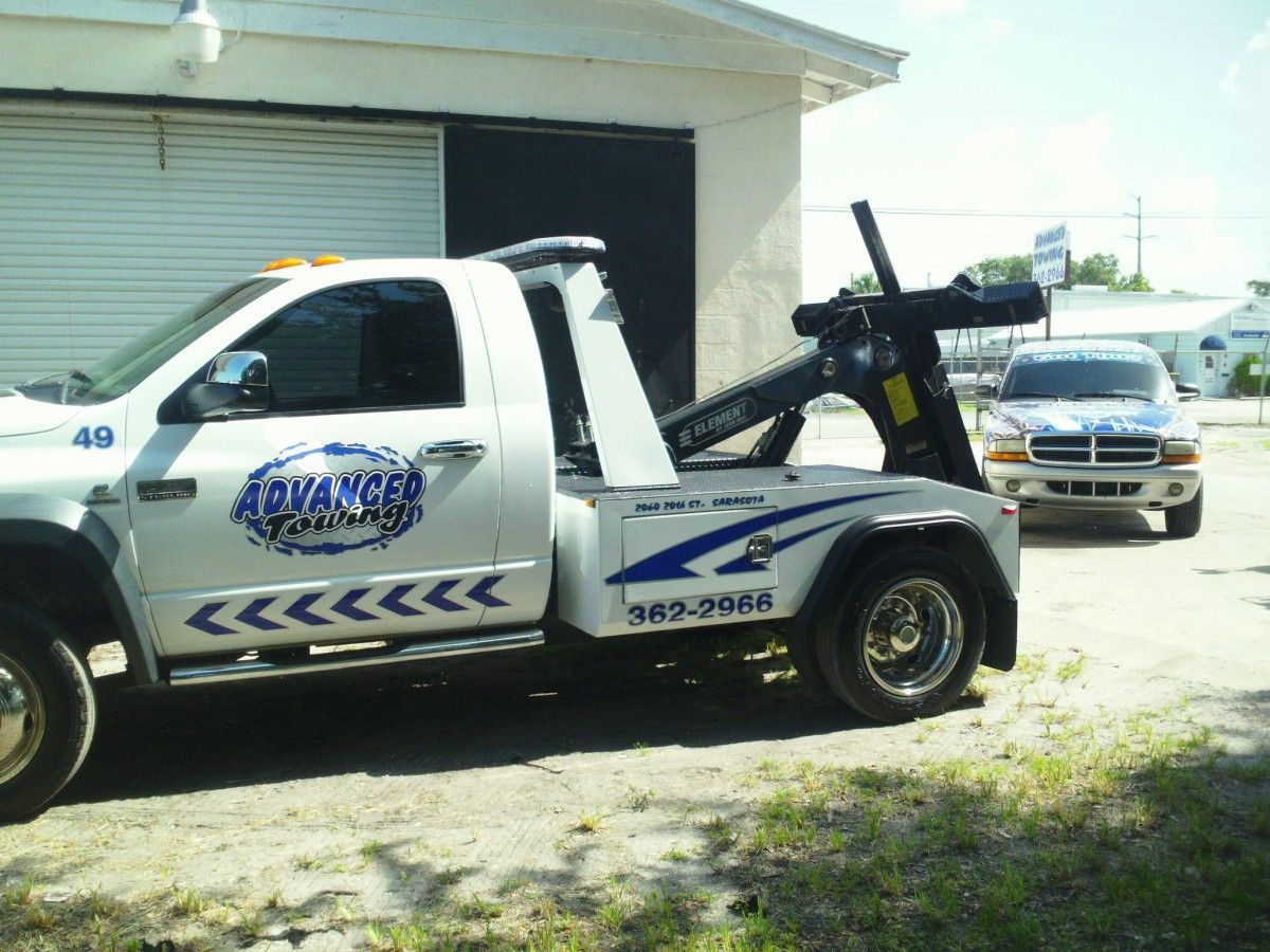 Tow truck parked beside a garage, with a sedan behind it on a sunny lot.