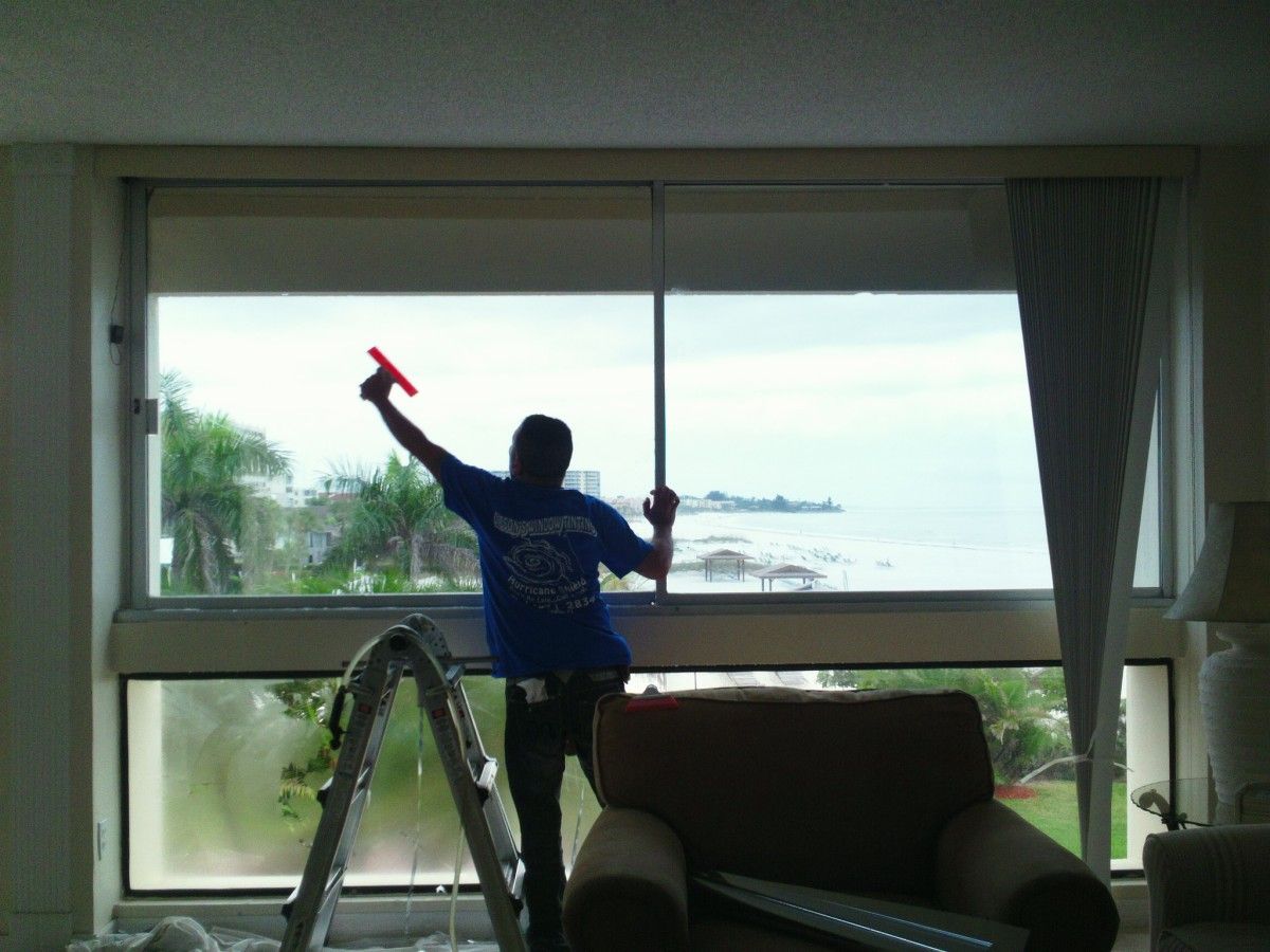 Person on a ladder painting the top of a large window with an ocean view.
