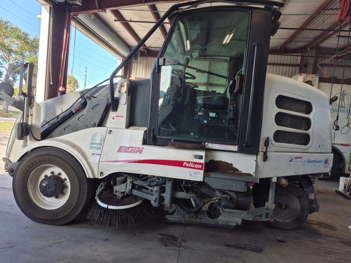 Damaged white street sweeper parked under a metal shed, side view with broken front components