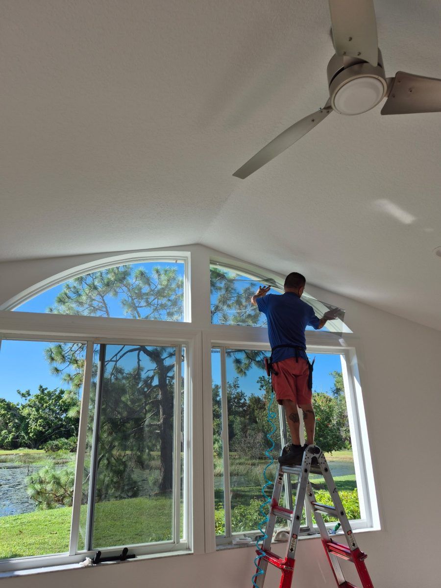 Person on a ladder painting the upper wall of a bright room with large windows and a ceiling fan.