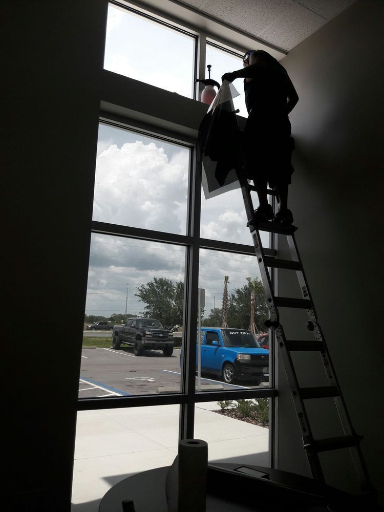 Worker on a ladder cleaning a high window inside a lobby with large glass panes.