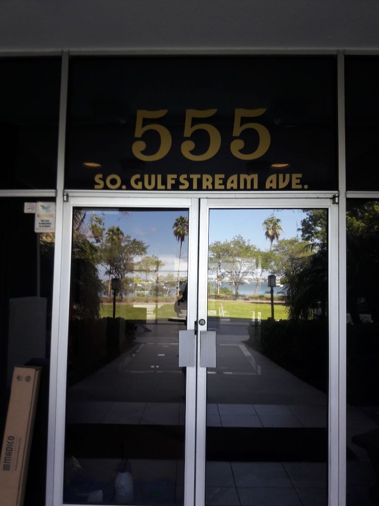 Glass entrance with “555” and “50 Gulfstream Ave.” above the doors, reflecting palm trees outside