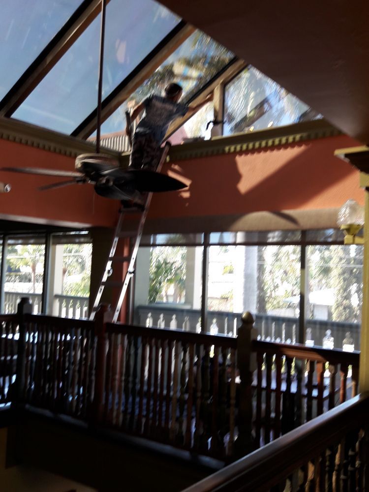 Worker on a ladder cleaning a skylight in a sunlit indoor space with wood railings and large windows