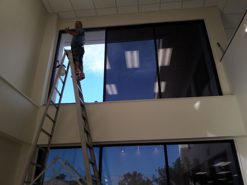 Worker on a ladder installing something near a large interior window in a building lobby.