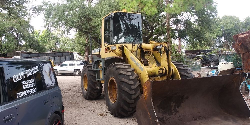 Yellow front loader with a raised bucket parked on a dirt lot near a black SUV and trees