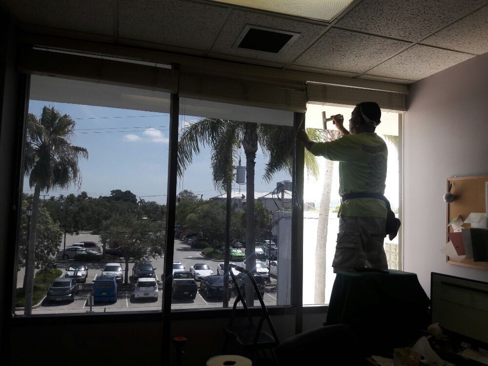 Person installing a window blind in an office, with parking lot and palm trees outside.