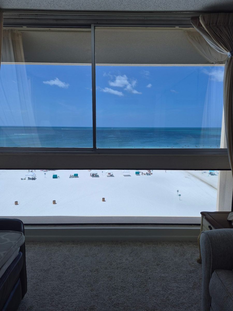 Ocean view through large window from a hotel room, with white sand and blue sky beyond.