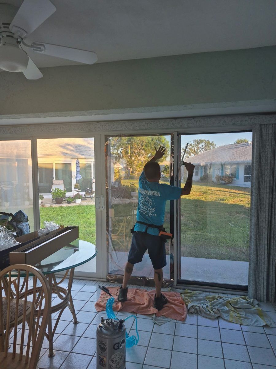Person cleaning a sliding glass door in a screened patio, holding a squeegee beside a bucket and cloths