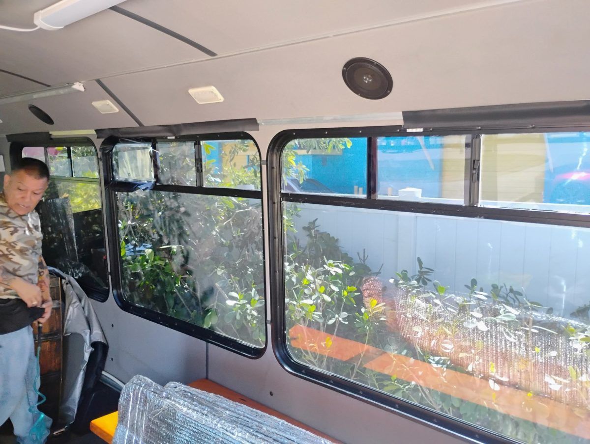 Interior of a van with large windows showing potted plants outside; a person stands at left.
