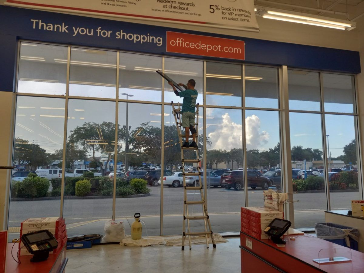 Worker on a ladder cleaning a storefront window beneath a “Thank you for shopping” sign.