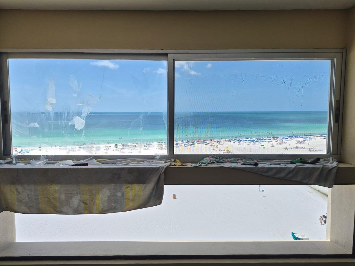 View through a window to a sunny beach and blue ocean, with curtains hanging below the sill