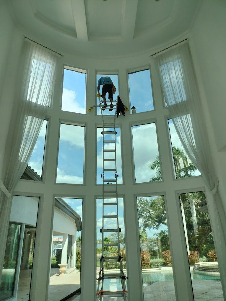 Worker on a ladder in a bright glass atrium with tall windows and a view of a pool outside