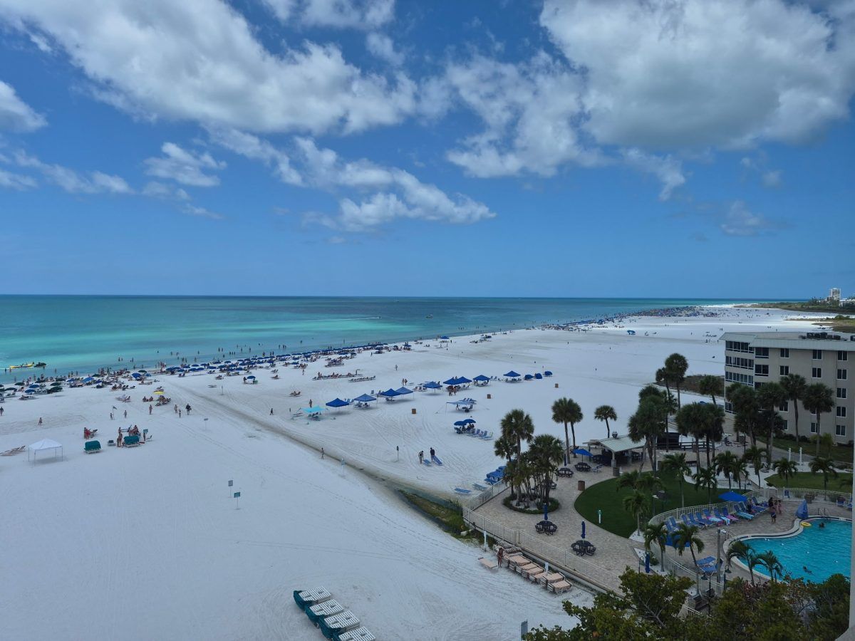 Sunny beach with turquoise water, white sand, umbrellas, and a resort pool under a partly cloudy sky