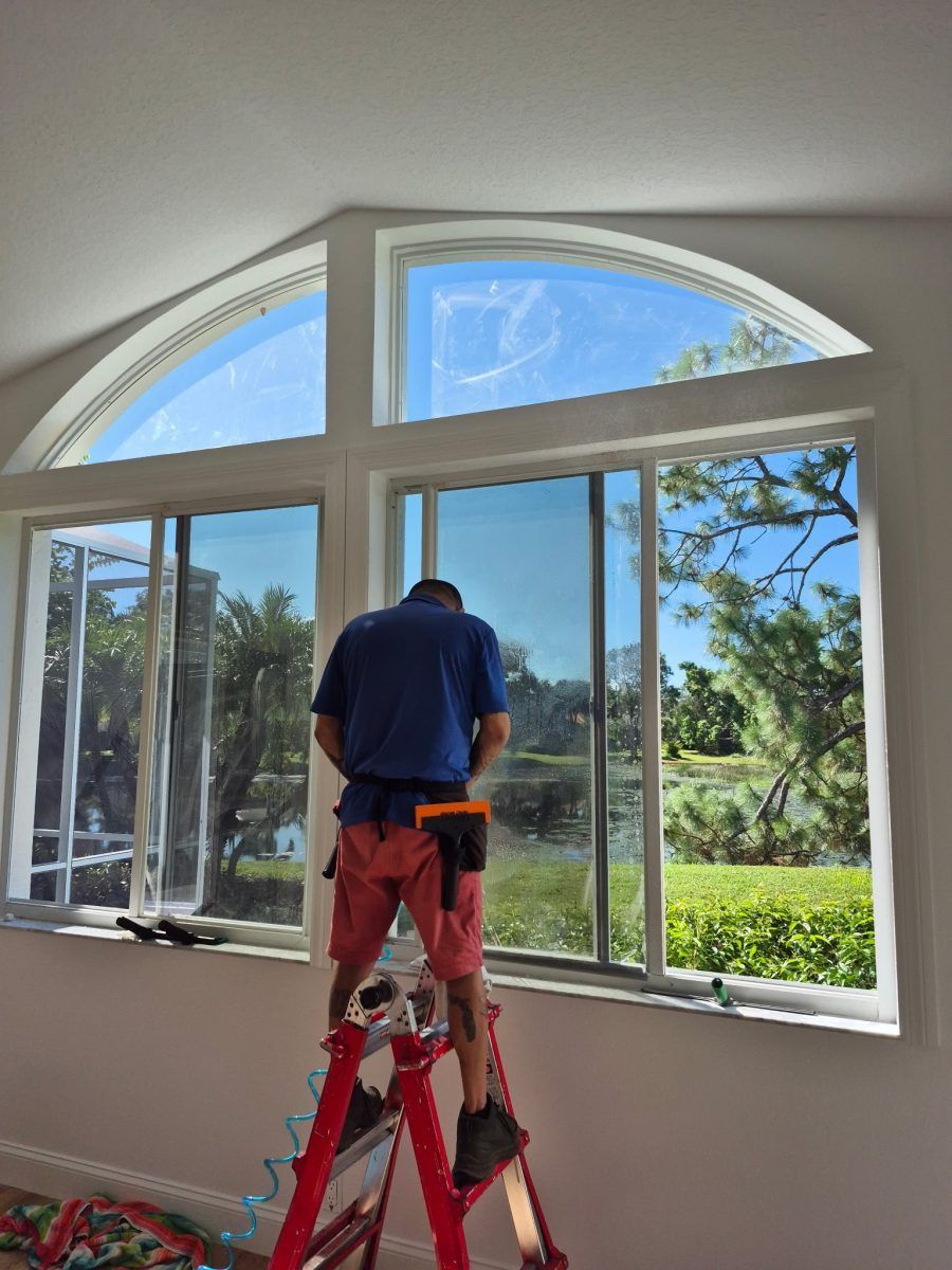 Worker on a red ladder installing a large window in a bright room with arched panes.