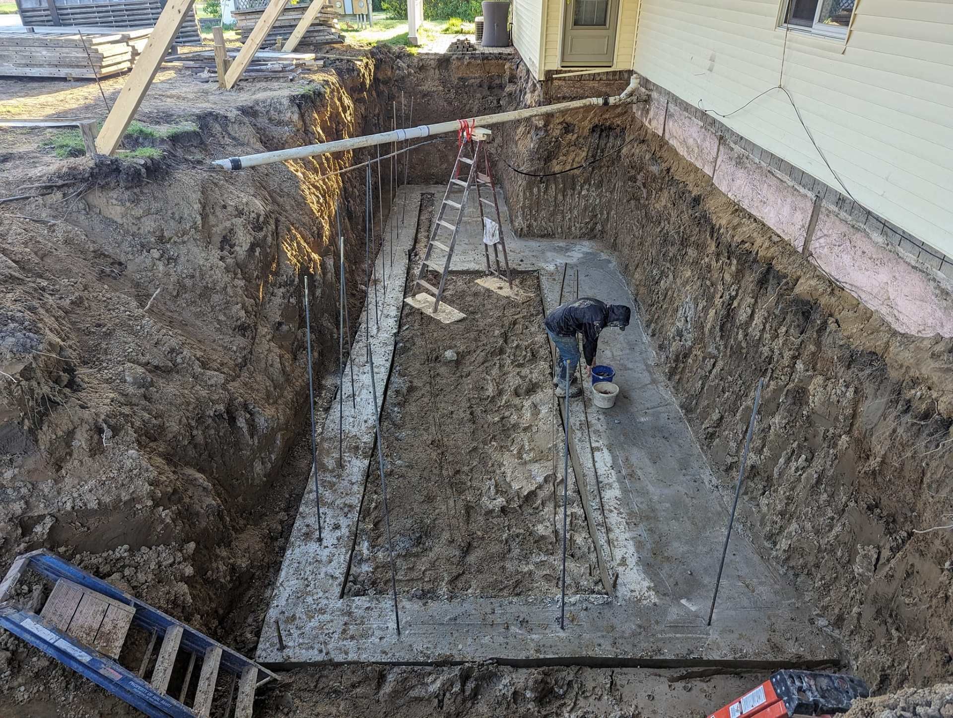 A worker in a construction trench beside a house, standing on a new concrete foundation with metal rebar stakes.