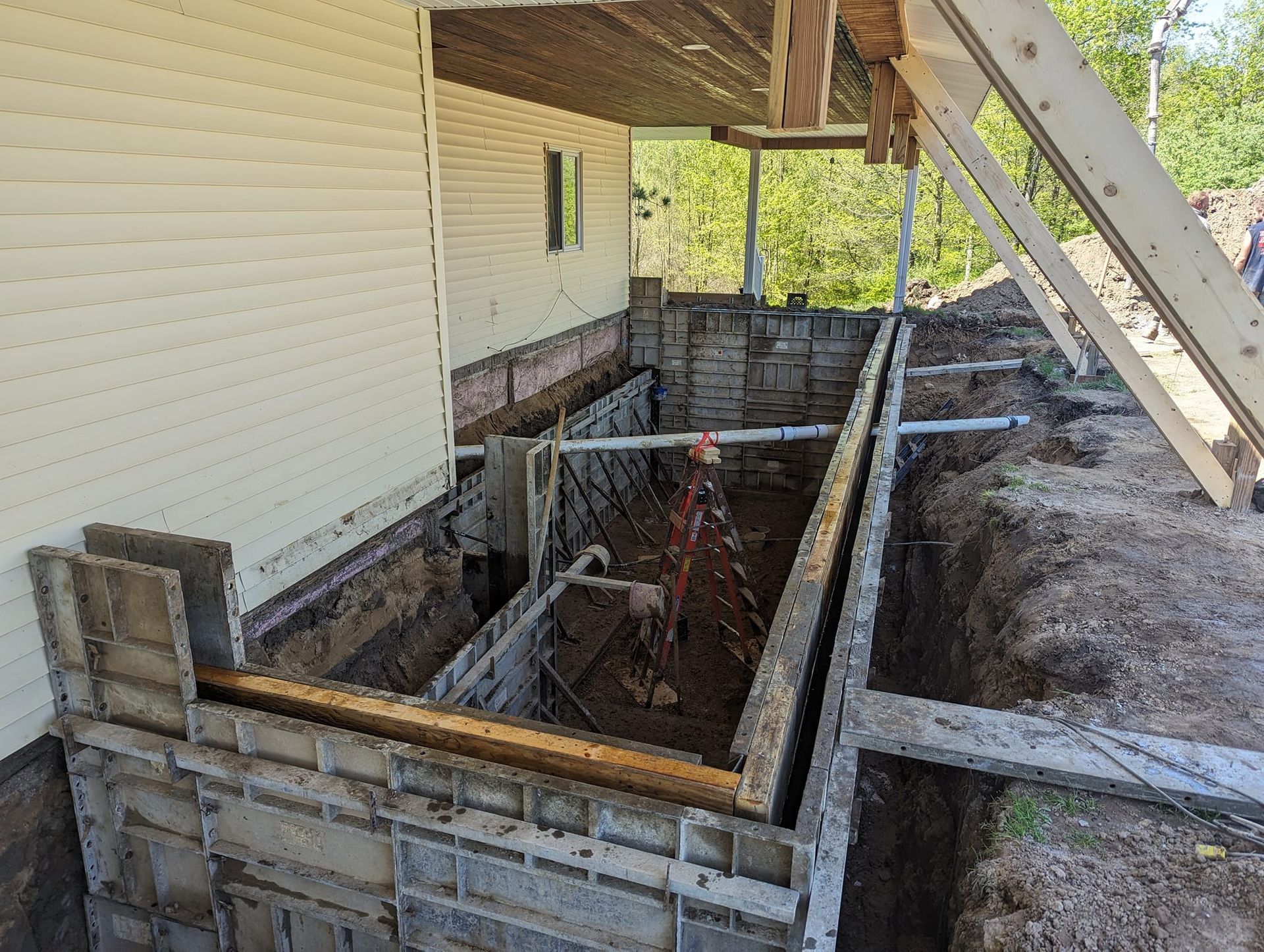 A construction site showing concrete foundation forms being installed for a home extension, with an open trench.