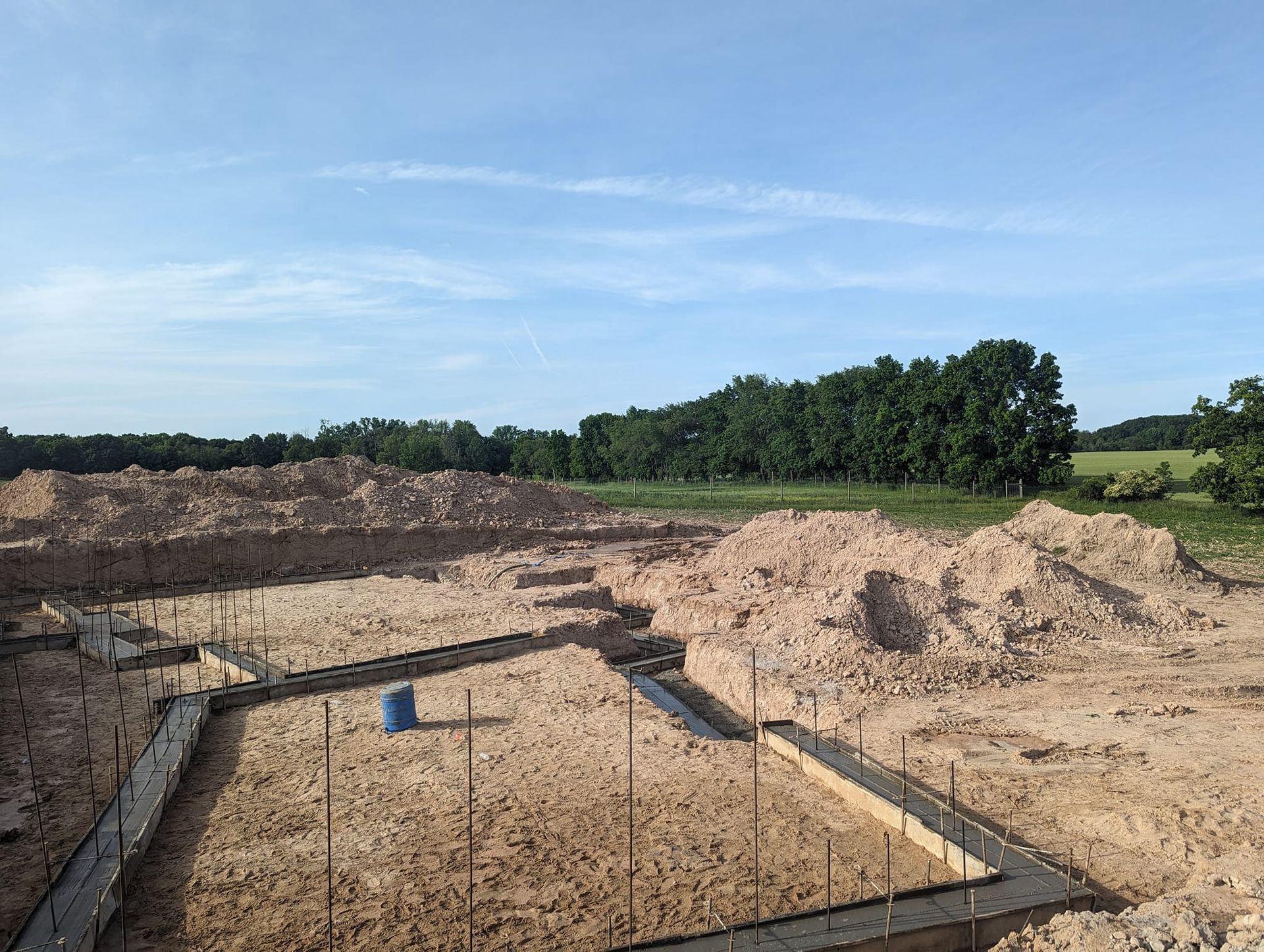 A construction site with concrete foundation footings poured in a sandy, excavated lot under a clear blue sky.