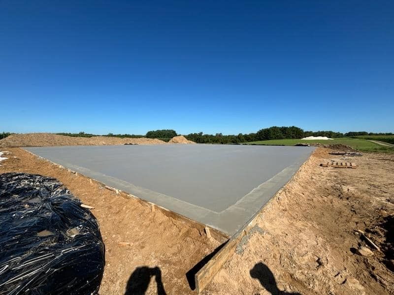 A large, smooth, freshly poured concrete slab sits in a dirt construction site under a clear blue sky.