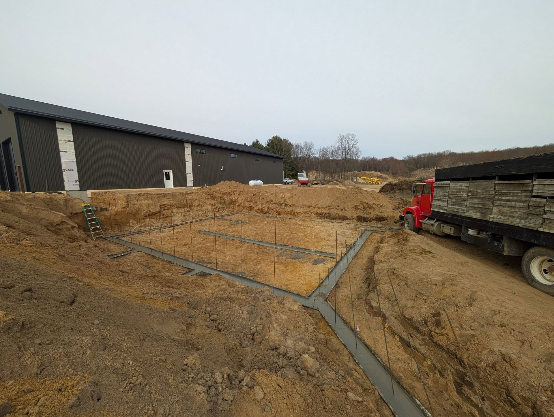 A construction site showing concrete footings poured in a rectangular trench next to a dark metal building and dump truck.