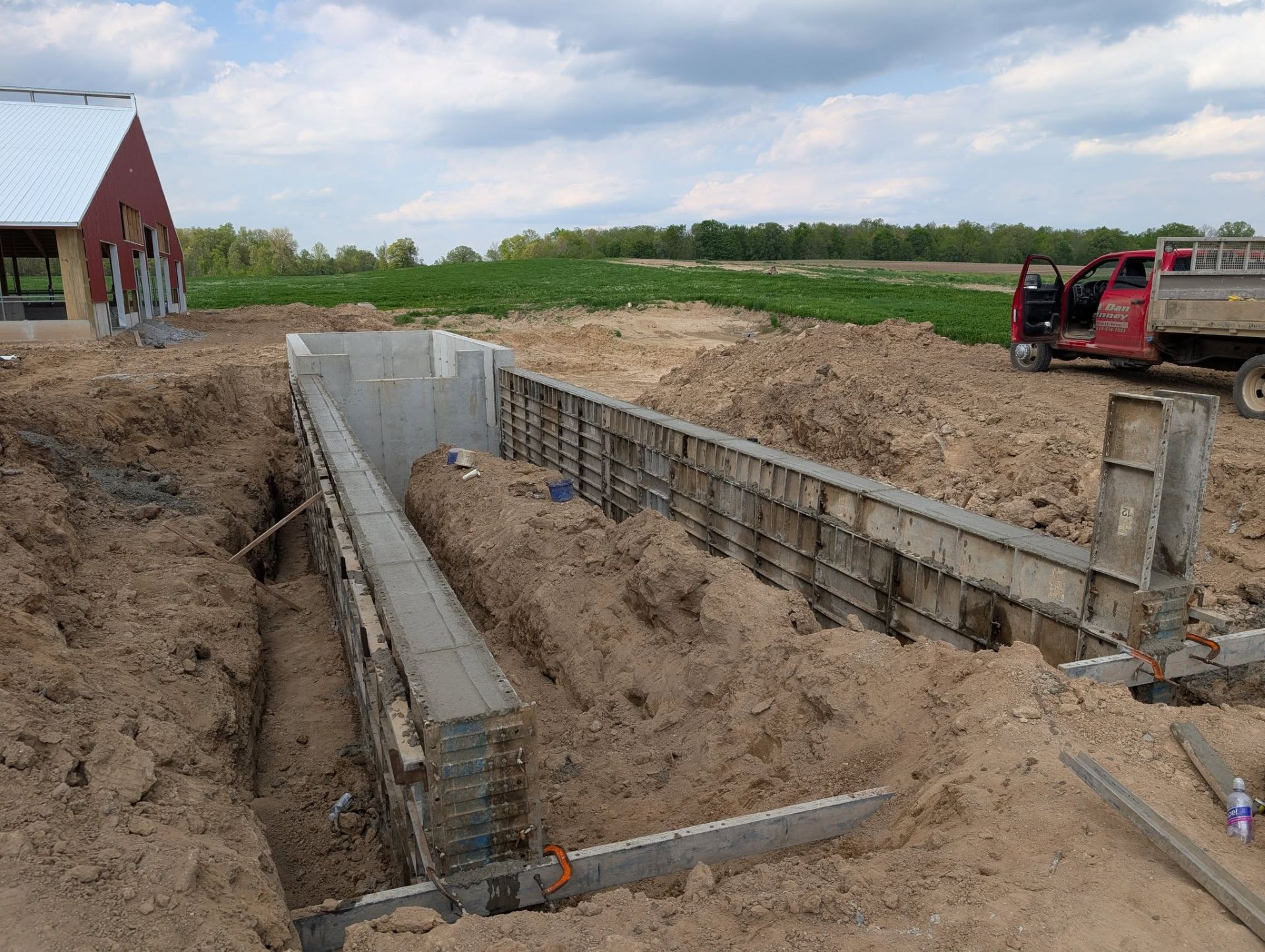 A construction site showing concrete wall forms installed in an excavated trench, with a red truck parked nearby.