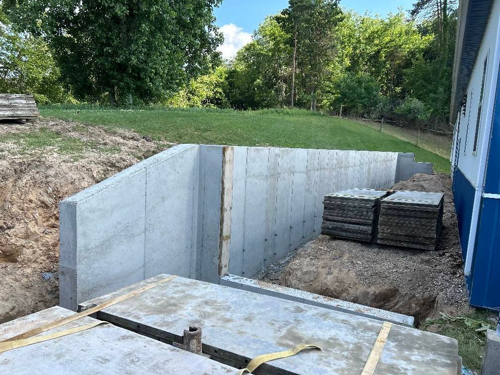 A newly installed gray concrete retaining wall sits in an excavated trench beside a building, with stacks of metal forms.