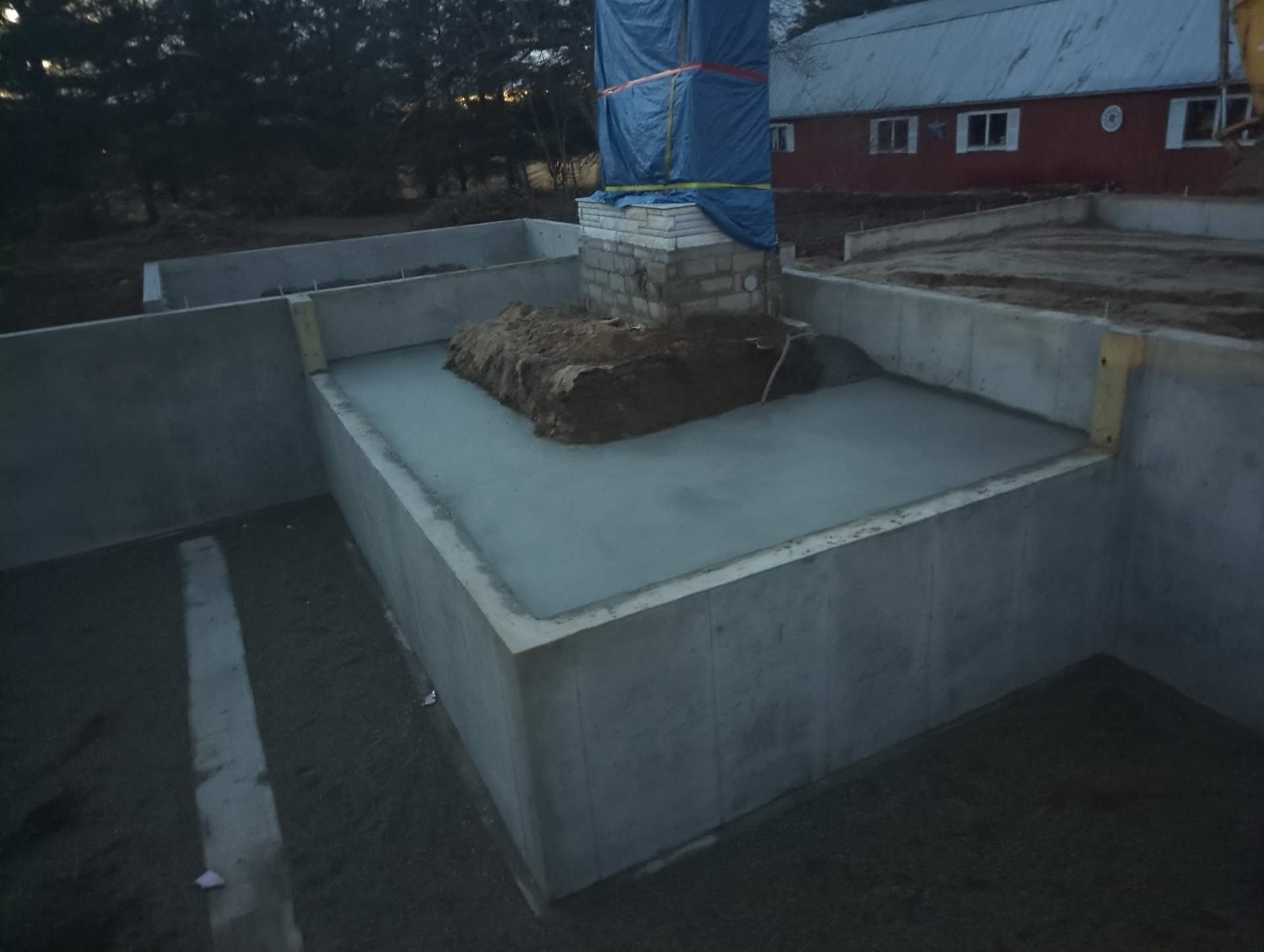Newly poured concrete slab foundation surrounding a brick chimney base covered in blue plastic on a construction site.