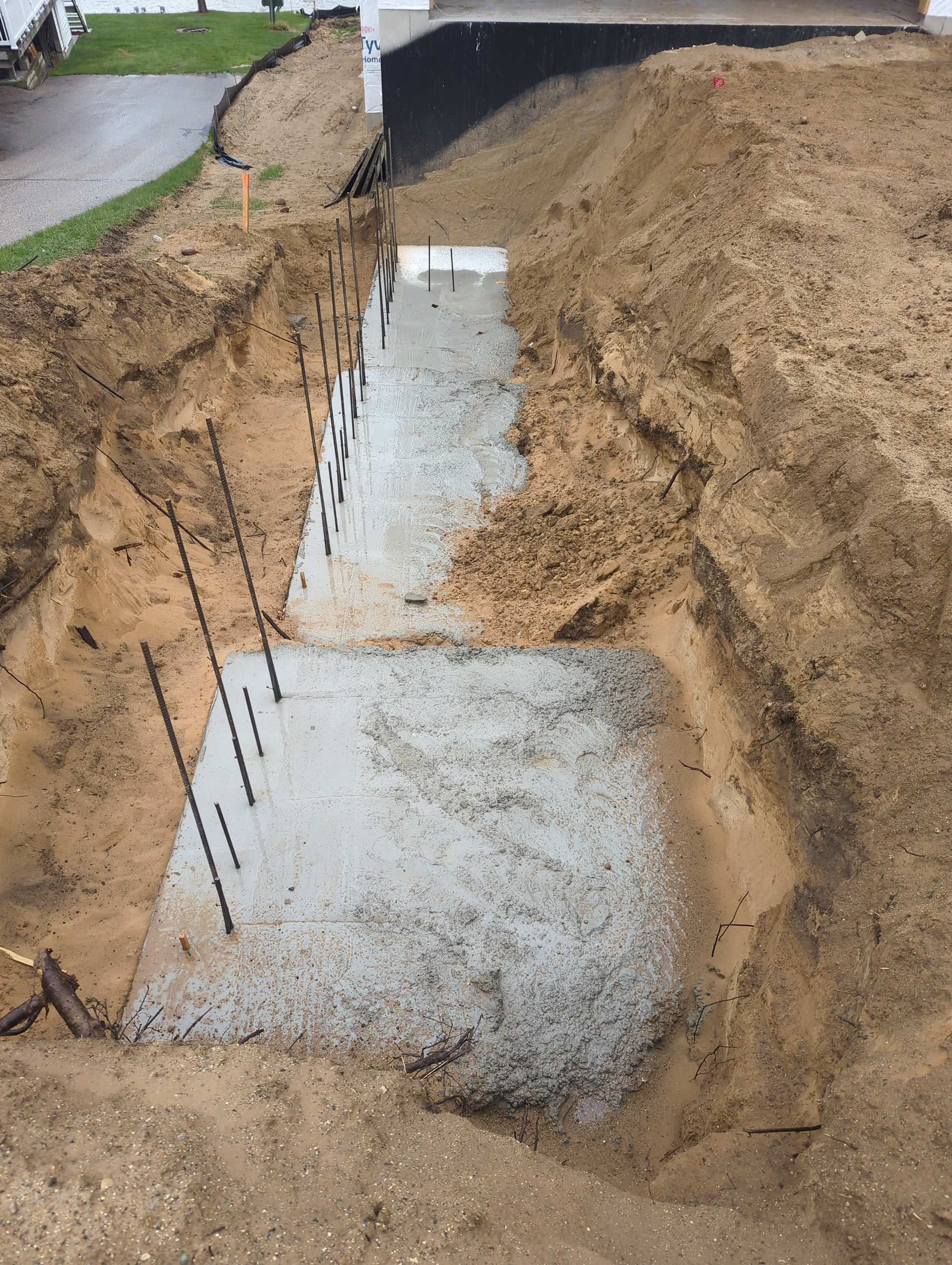 A newly poured concrete foundation slab sits in a deep, sandy dirt trench beside a building wall with black waterproofing.