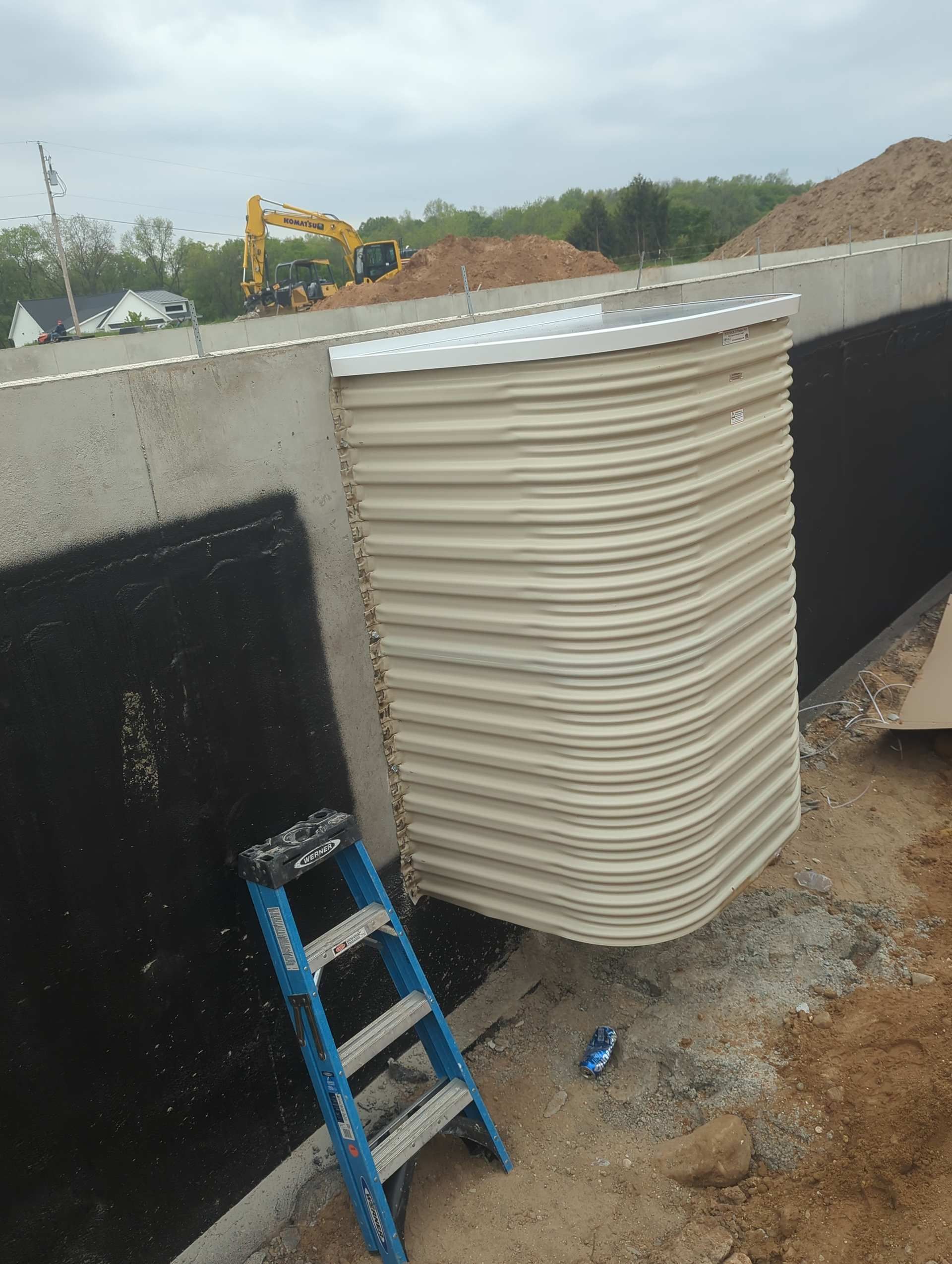 A beige corrugated window well attached to a concrete foundation wall with a blue ladder resting nearby at a job site.
