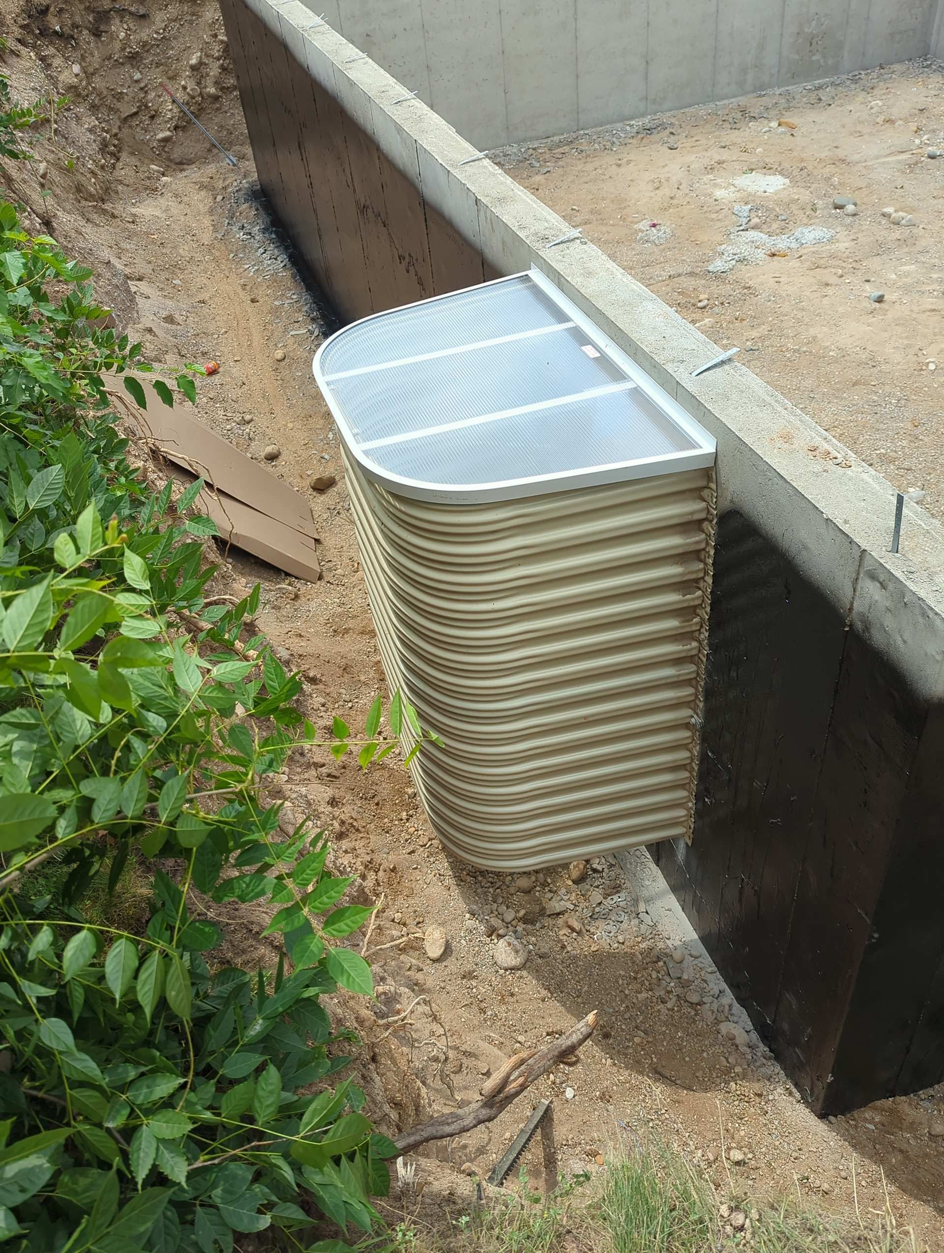 A tan, corrugated basement window well with a metal grate cover installed against a concrete foundation wall.
