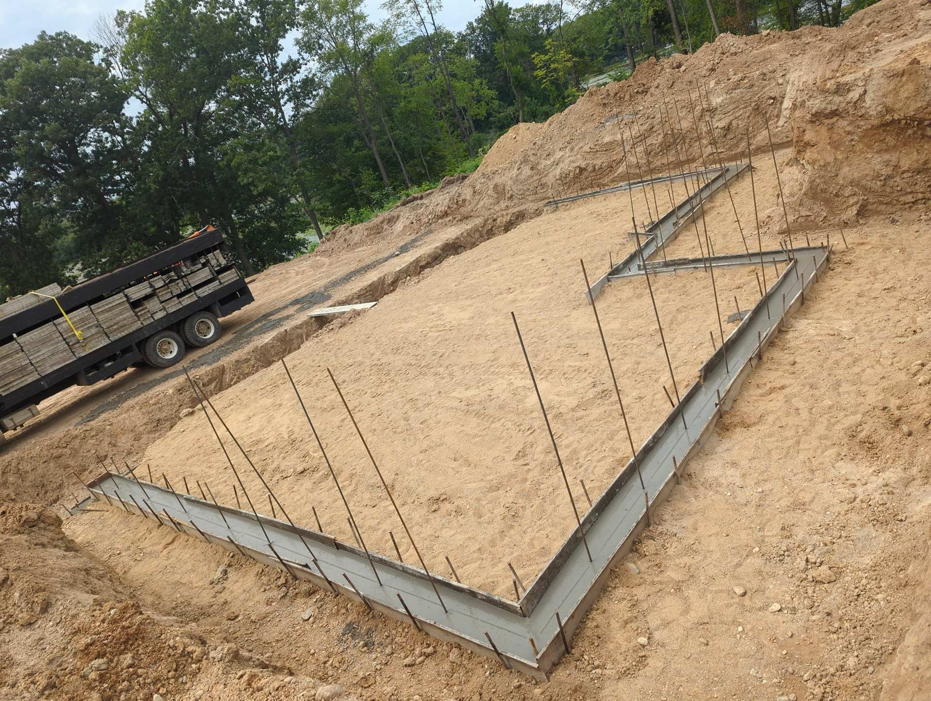 A concrete house foundation with vertical rebar stakes rising from the perimeter footings on a dirt lot.