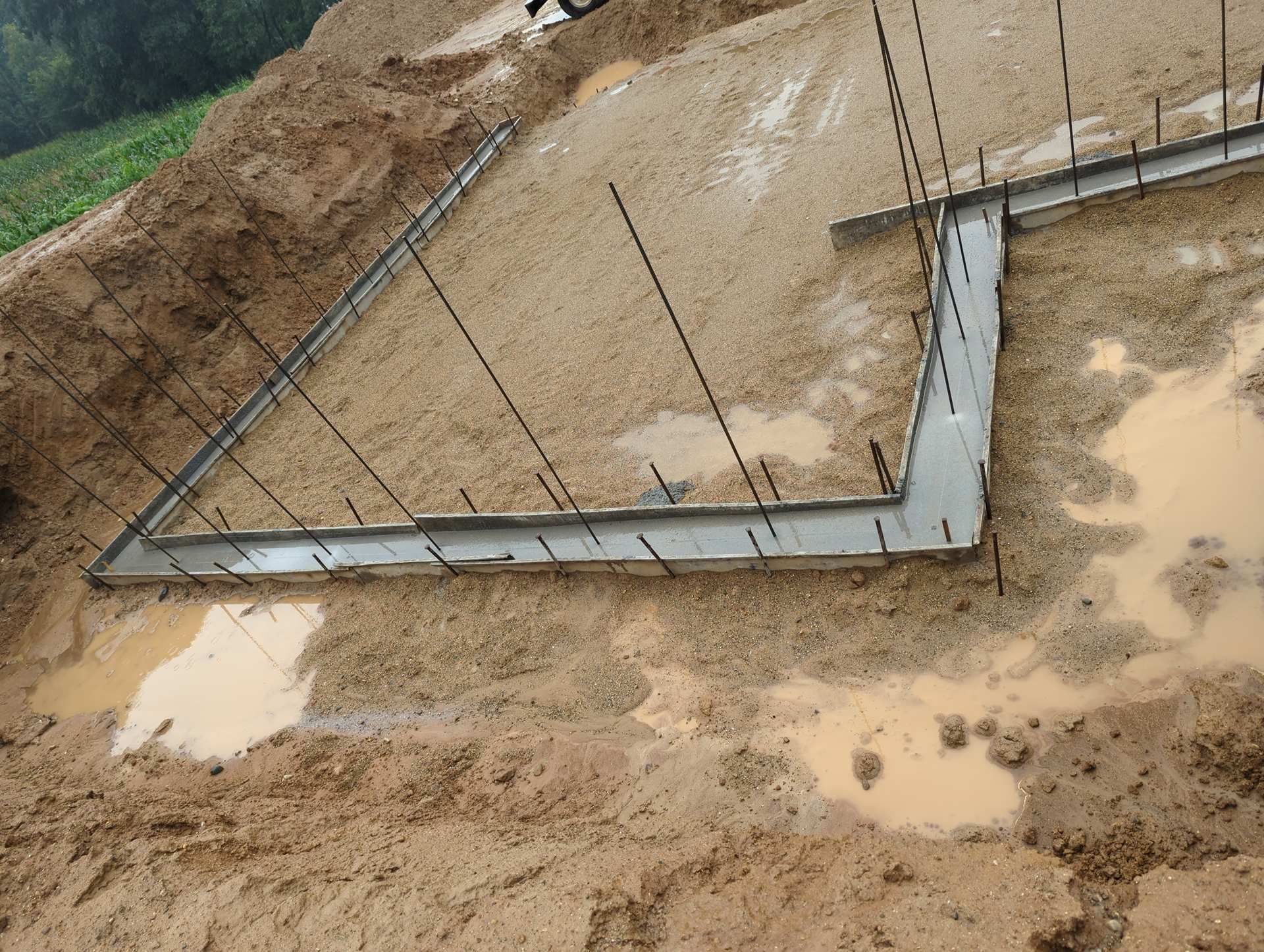 Concrete foundation footings with vertical rebar uprights in a muddy, excavated dirt site.