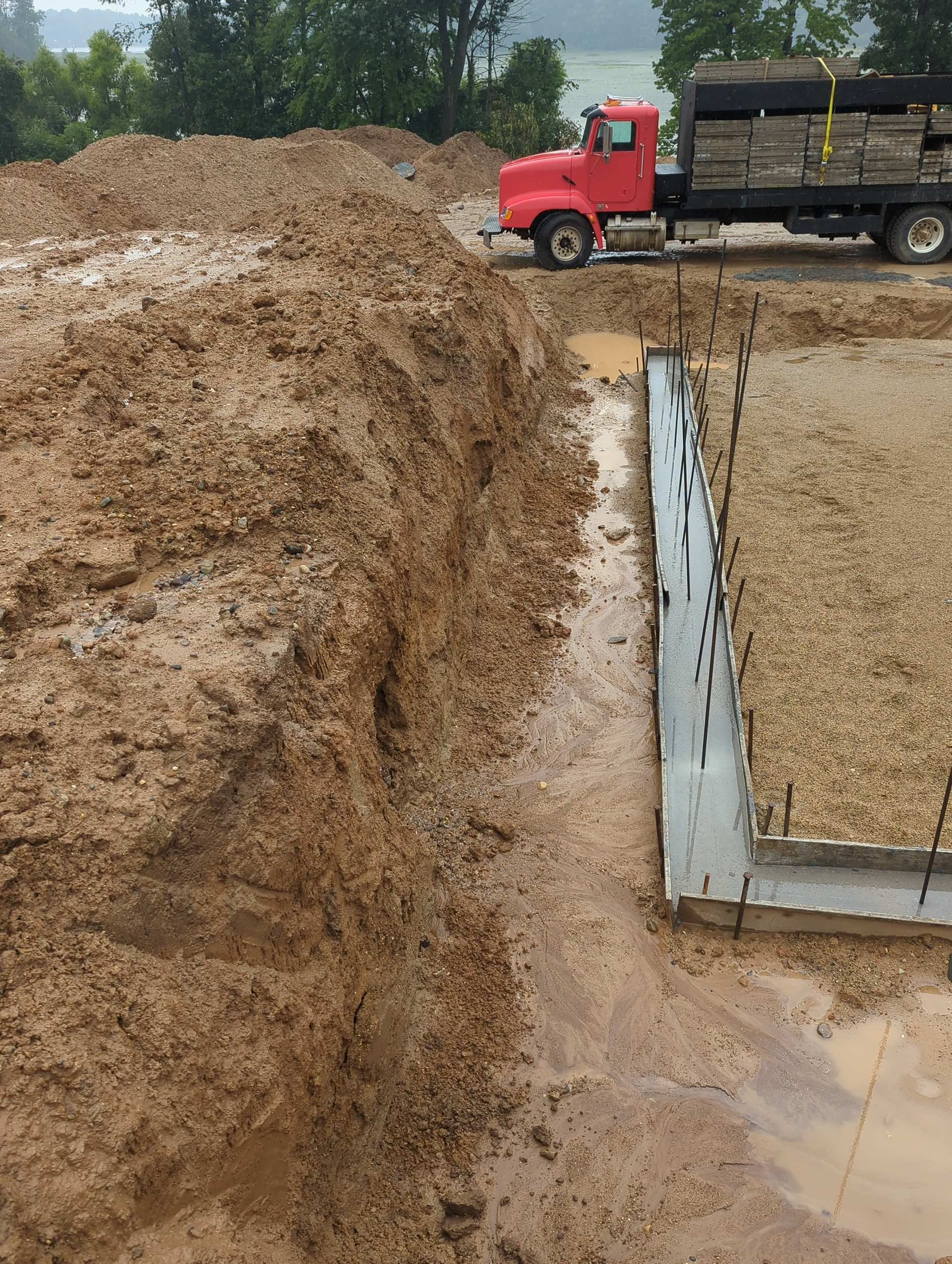 A red truck parked near a dirt excavation site next to a concrete foundation footer for a building.