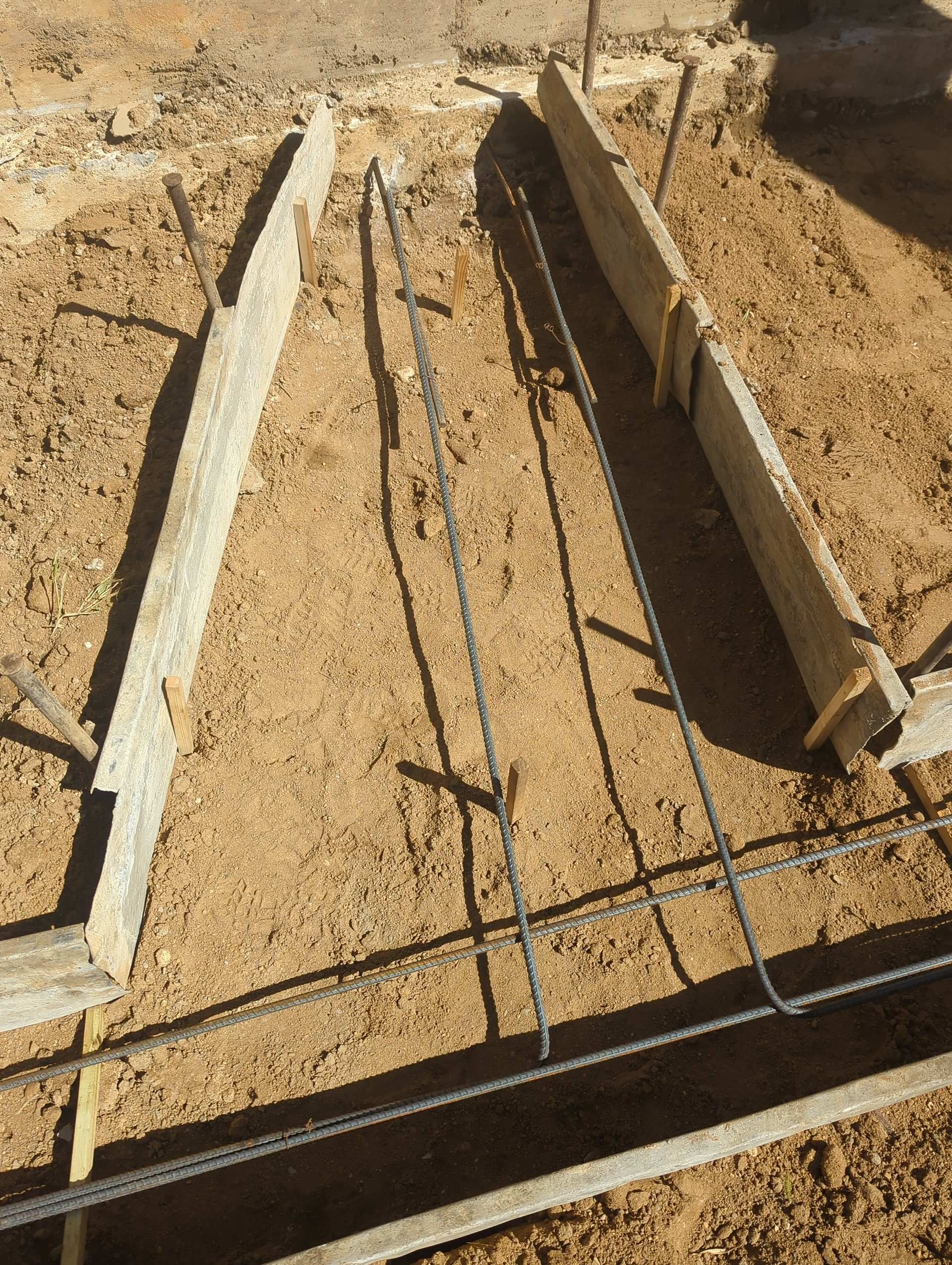 A construction site showing wooden formwork boards for a concrete foundation, with steel rebar laid out on the dirt.