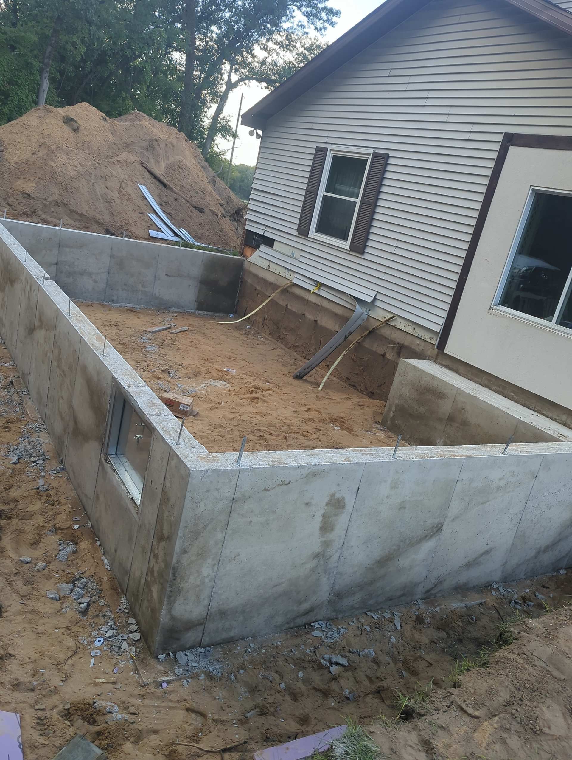 A concrete foundation for a room addition under construction next to a house with beige siding.