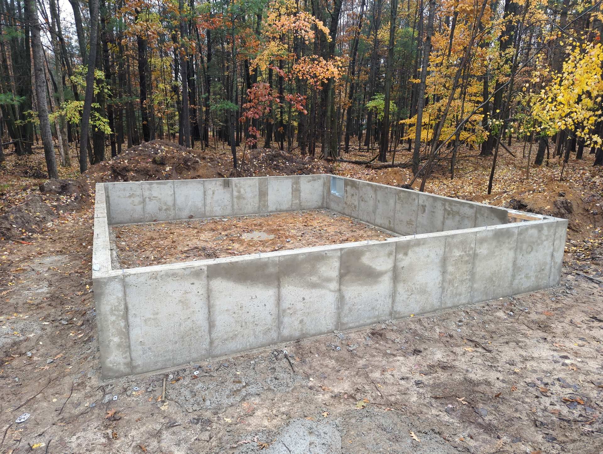 A rectangular concrete foundation for a small structure sits in a wooded area filled with fallen autumn leaves.