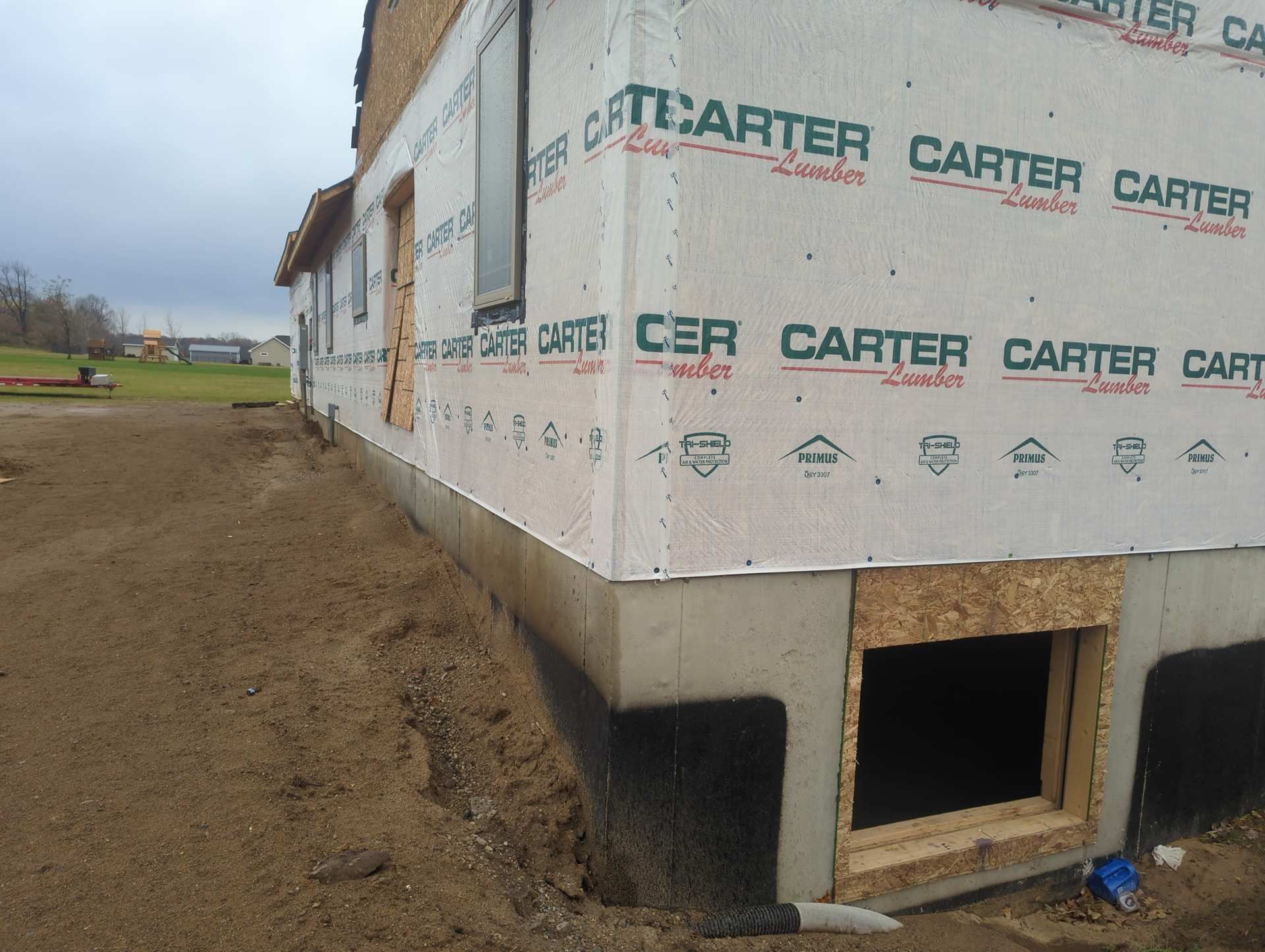 Exterior view of a residential construction site featuring a concrete foundation and house wrap on a gravel lot.