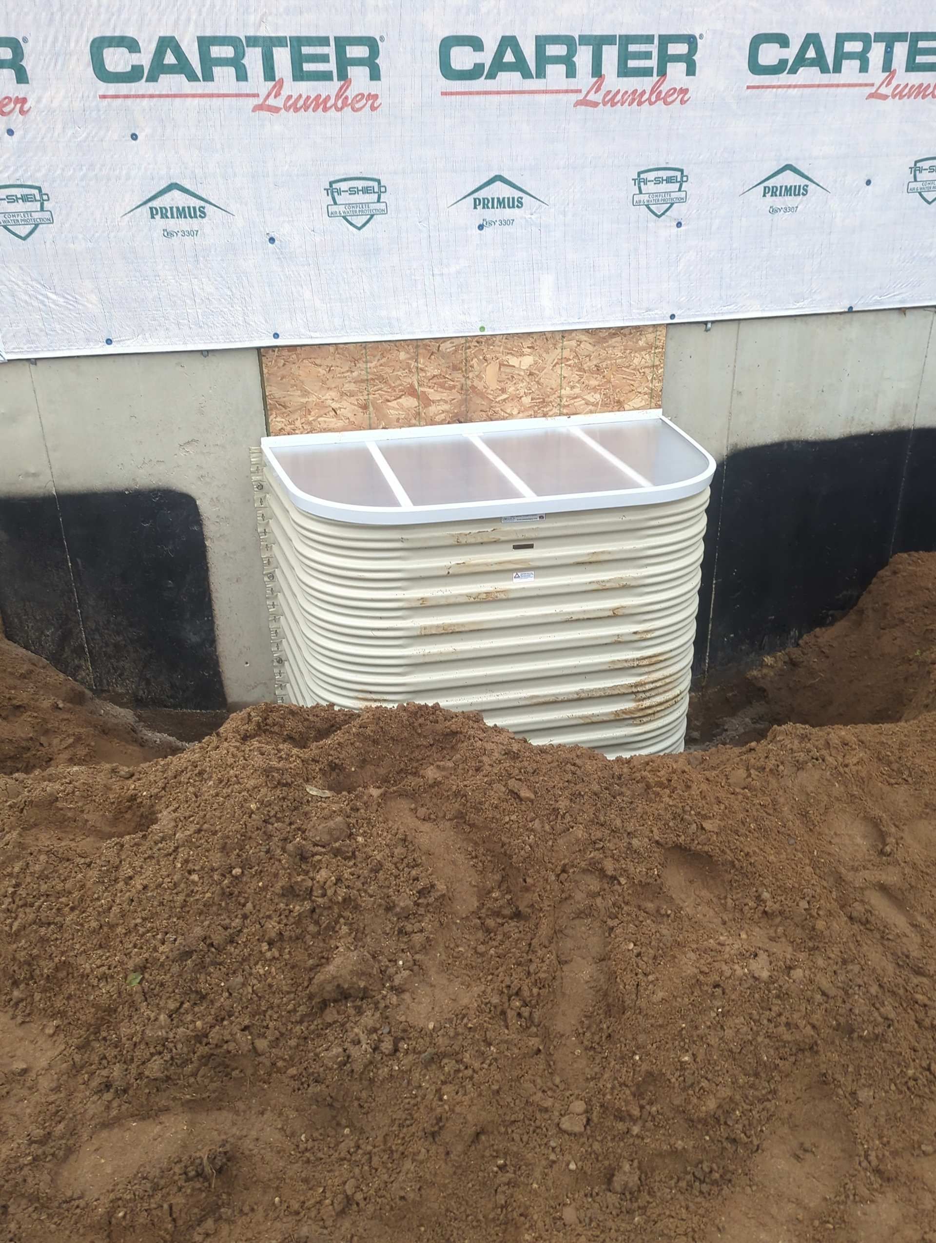 A white corrugated window well installed against a concrete foundation wall with soil piled in the foreground.