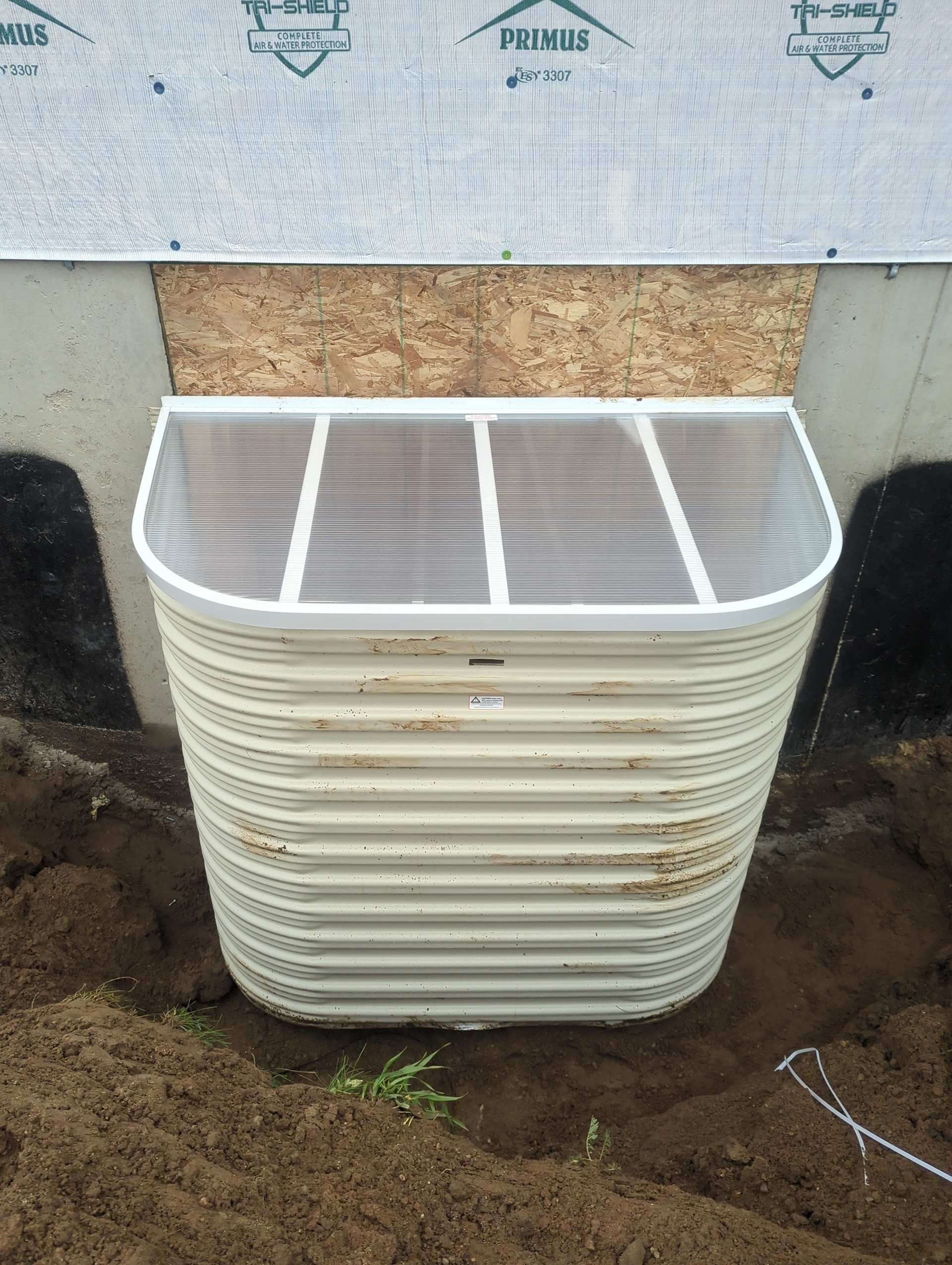 A corrugated metal window well with a clear plastic cover installed against a basement foundation wall.