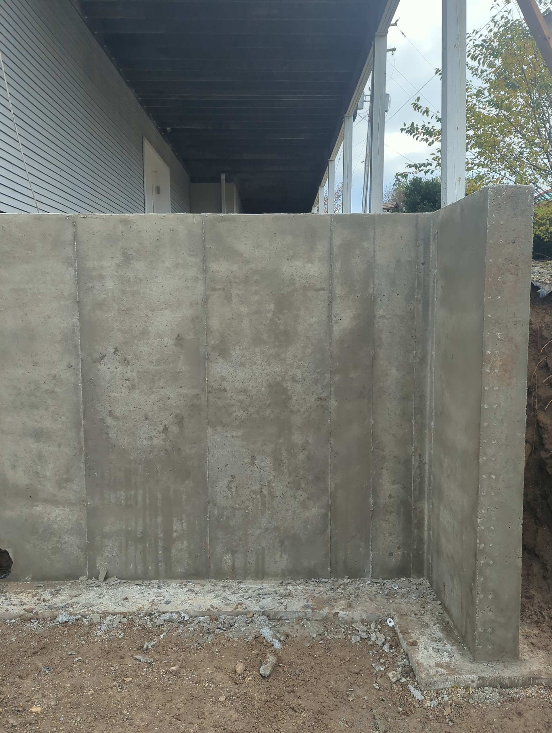 A newly poured concrete retaining wall stands next to a building, with gravel and dirt at its base.